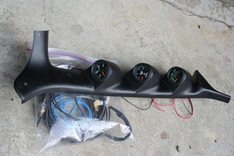 Black plastic gauge pod with three gauges and wiring harness, lying on concrete