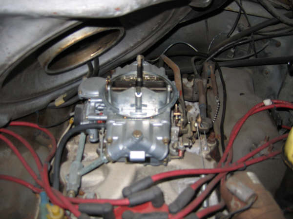 Four-barrel carburetor on intake manifold with red plug wires and throttle linkage, air cleaner removed