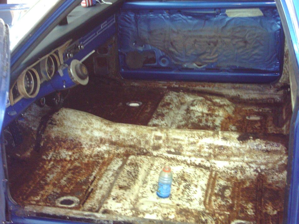 Stripped car interior with rusty bare floor pan, blue dash and door panel, and a Gatorade bottle