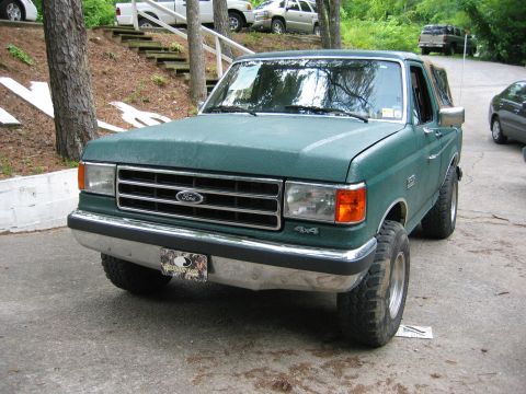 1990 Ford Bronco - A green 1990 Ford Bronco is parked on a paved surface, with other vehicles visible in the background.