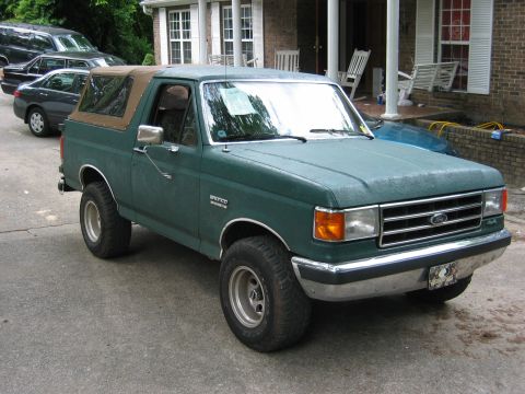 1990 Ford Bronco - This Ford Bronco is parked in front of a brick house with other vehicles nearby.