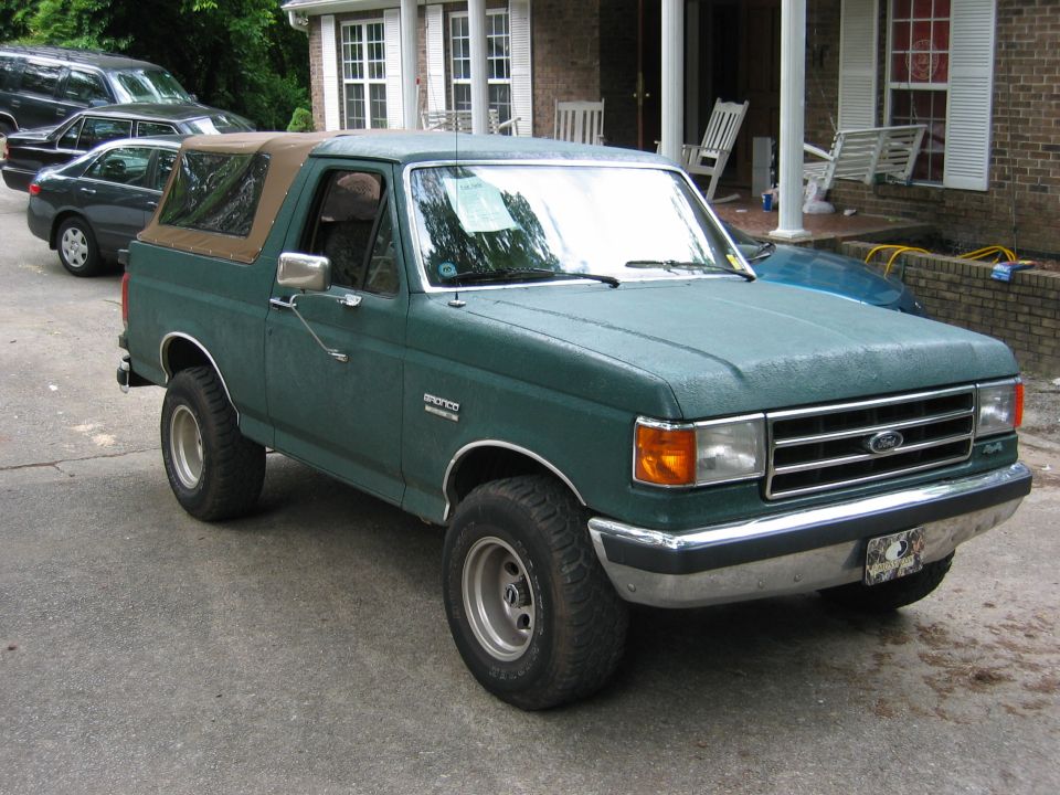 Green Ford Bronco with tan soft top parked in driveway. Other cars and a house in background