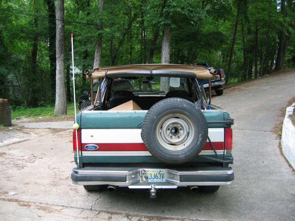 Rear view of a green and red Ford Bronco with a spare tire mounted on the back. Another vehicle is visible behind it