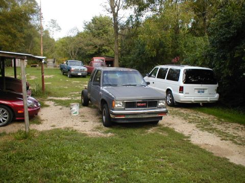 1998 Ford Taurus - A gray GMC pickup truck and a white minivan are parked outdoors on a dirt path.