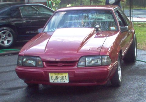 1988 Ford Bronco - A red coupe with a prominent aftermarket hood is parked outside next to another vehicle.