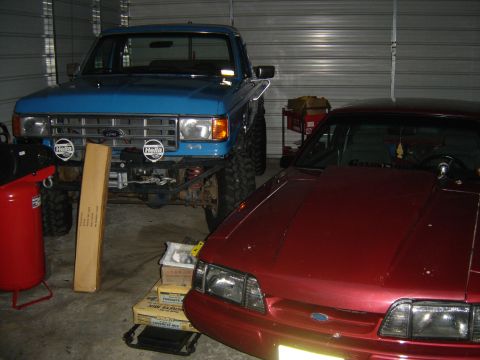 1988 Ford Bronco - Two vehicles, a blue pickup and a red coupe, are parked side-by-side inside a garage.