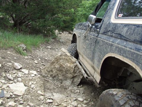 1986 Ford Bronco - A Ford Bronco maneuvers over a rough, rocky terrain with dense foliage in the background.