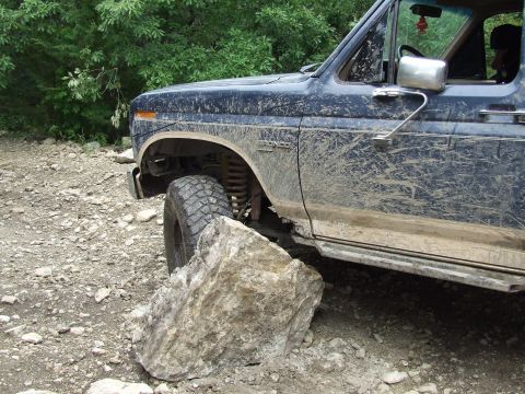 1986 Ford Bronco - A muddy Ford Bronco navigates a rocky trail, its front tire positioned near a large boulder.