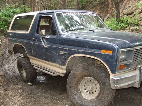 1986 Ford Bronco - Muddy Ford Bronco navigating off-road terrain with lifted suspension and large tires.
