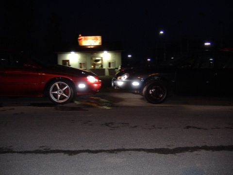 1998 Ford Taurus - Two cars, a red 1998 Ford Taurus and a dark vehicle, are parked nose-to-nose in front of a lit building at night.
