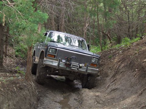 1986 Ford Bronco - This Ford Bronco navigates a challenging muddy path in a wooded area, with a "BRONCO.COM" banner on its windshield.