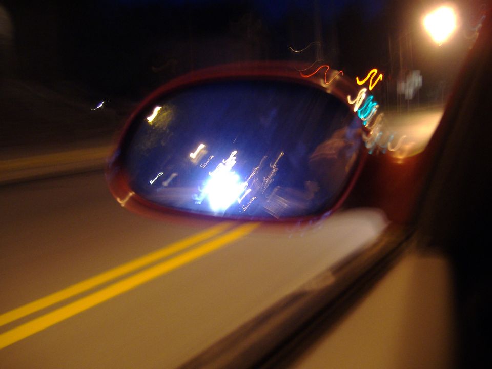Side mirror reflecting bright lights at night, with double yellow lines on the road