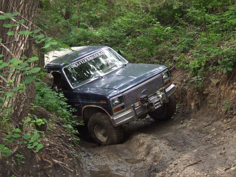 1986 Ford Bronco - This dark blue SUV navigates a challenging muddy off-road trail, its tires struggling for traction.