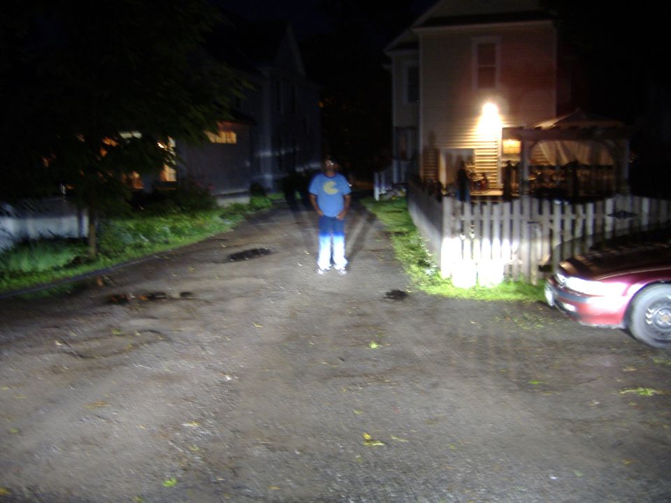 Man in blue shirt and jeans stands in a dark driveway with houses and a car visible