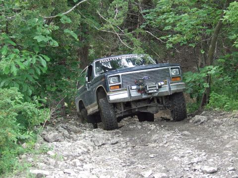 1986 Ford Bronco - A dark blue Ford Bronco navigates a challenging, rocky, wooded path.