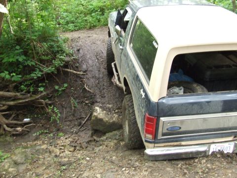 1986 Ford Bronco - Ford Bronco navigates a challenging muddy path with dense foliage on the side.