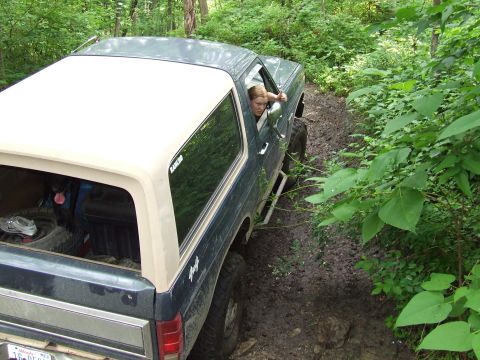 1986 Ford Bronco - A woman drives a classic Ford Bronco through a wooded, muddy off-road path.