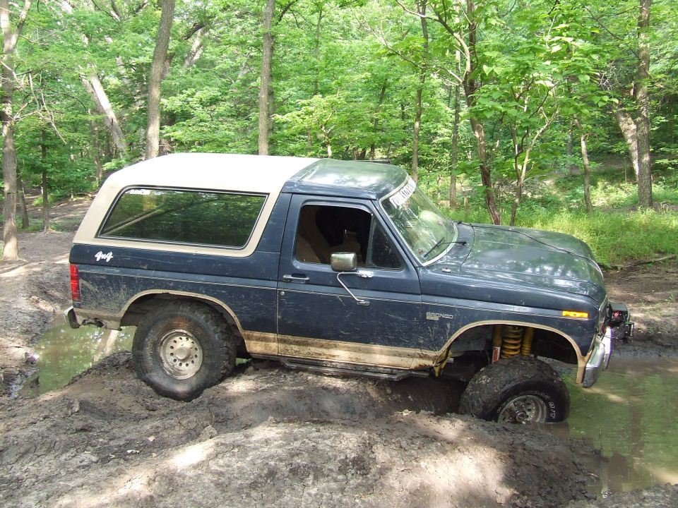 Dark blue Ford Bronco with a white top is driving through mud and water in a wooded area. It has a 4x4 decal on the rear quarter panel