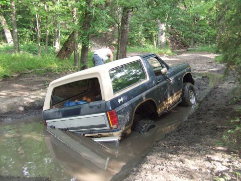 1986 Ford Bronco - A 1986 Ford Bronco is stuck in a muddy trail, with its rear wheels submerged in water and mud.