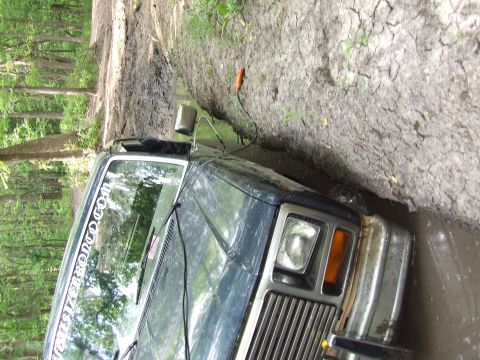 1986 Ford Bronco - A dark blue SUV is stuck in a muddy ditch, with trees visible on the left side of the frame.