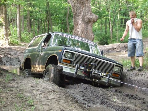 1986 Ford Bronco - A 1986 Ford Bronco is stuck in a muddy ditch while a man attempts to pull it out with a rope.