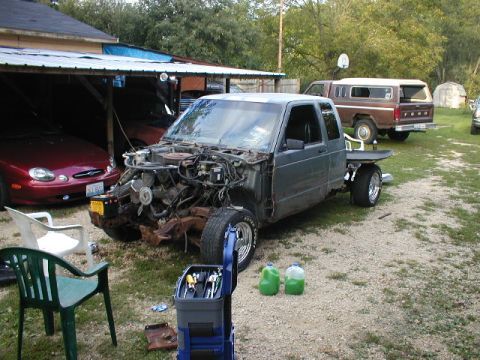 1998 Ford Taurus - A gray pickup truck is missing its front end, parked in a driveway with a red car and a brown camper van.