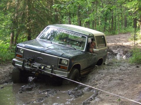 1986 Ford Bronco - A Ford Bronco navigates a difficult muddy off-road path, with a person visible inside the vehicle.