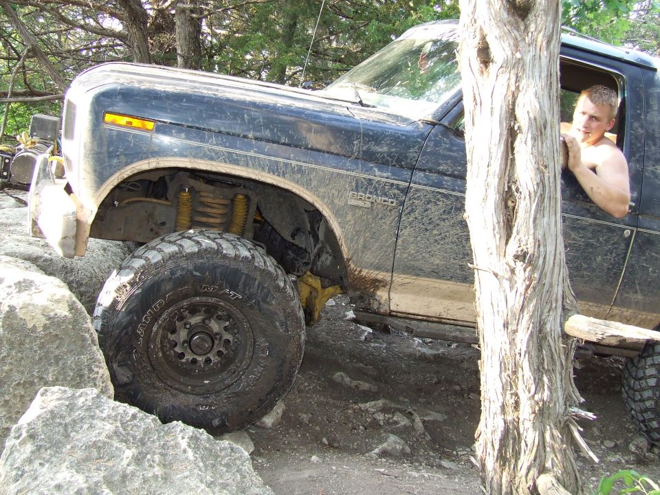 Mud-covered 1980s Ford Bronco navigating rocky terrain, driver visible through window
