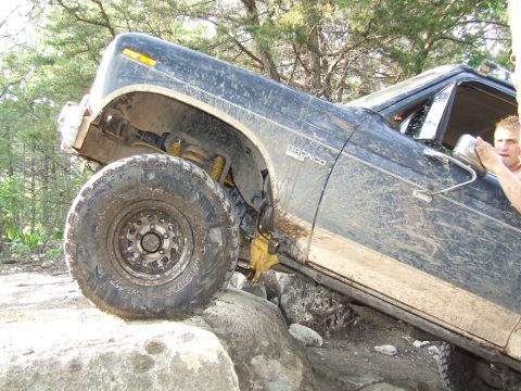 1986 Ford Bronco - A dark blue Ford Bronco navigates a rocky trail, showing significant mud coverage and off-road modifications.