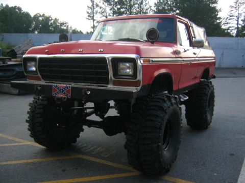 1979 Ford Bronco - A heavily modified red Ford Bronco sits on a paved lot, its lifted suspension and oversized off-road tires.