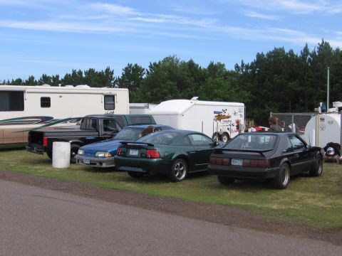 1989 Ford Mustang - A black 1989 Ford Mustang coupe is parked on the grass next to other vehicles and trailers.