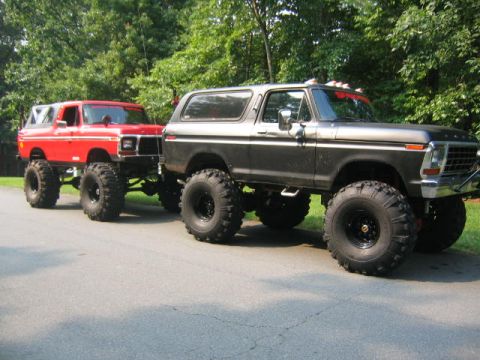 1979 Ford Bronco - Two lifted first-generation Ford Bronco SUVs parked on a paved driveway surrounded by trees; a red one behind and a gray/primer-colored one…