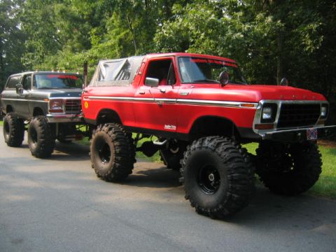 1979 Ford Bronco - A bright red late-1970s full-size Ford Bronco sits high on a heavy lift with black wheels and huge mud-terrain tires in a driveway, its whi…