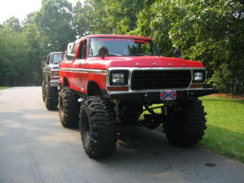 1979 Ford Bronco - Three-quarter front view of a heavily lifted red and silver 1978-79 Ford Bronco on huge mud tires, parked on a tree-lined road with a roll-…