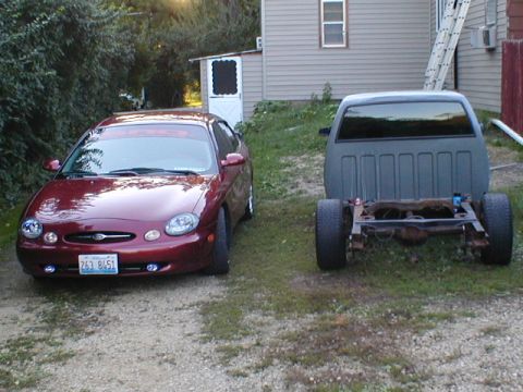 1998 Ford Taurus - A red 1998 Ford Taurus is parked on a gravel driveway next to a bare truck frame.