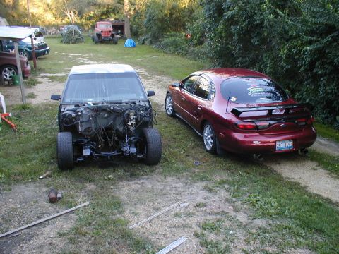 1998 Ford Taurus - A red 1998 Ford Taurus is parked next to a disassembled vehicle. The Taurus has a spoiler and a custom license plate.