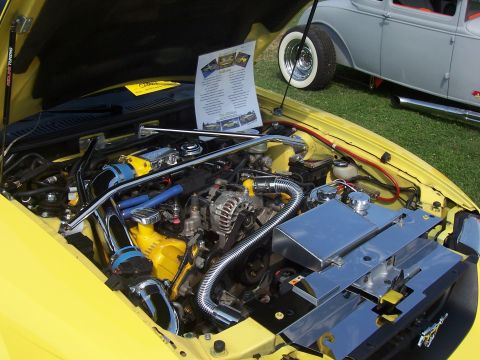 2002 Ford Mustang - Engine bay of a yellow 2002 Ford Mustang, custom chrome parts and a display sheet. A classic car is in the background.
