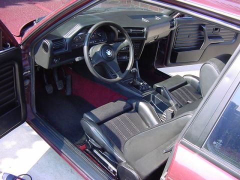 1984 BMW 3 Series - The interior of a 1984 BMW 3 Series features black seats with striped inserts, a red floor mat, and a BMW steering wheel.