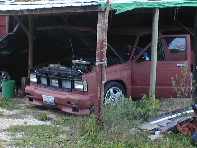 Red pickup truck with hood open, parked under a shelter. Another vehicle is partially visible to the left