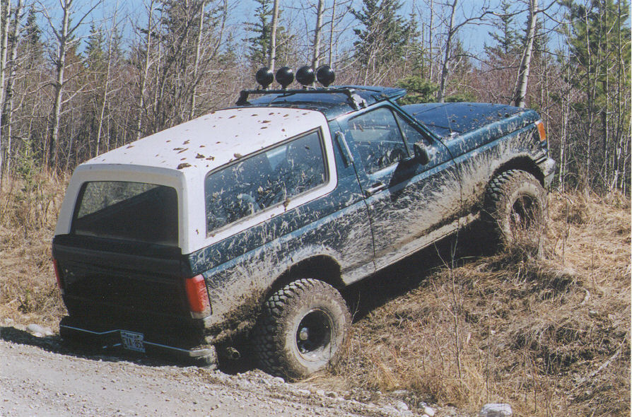 Muddy teal Ford Bronco with white top and four roof lights on a gravel trail