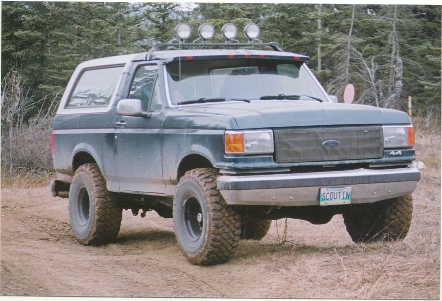Lifted green and white Ford Bronco on dirt trail with roof light bar and mud tires