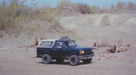 1988 Ford Bronco - A dark blue Ford Bronco with a white top is parked on a sandy trail with sparse vegetation.