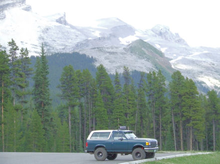 1988 Ford Bronco - A blue Ford Bronco is parked on a road with a backdrop of pine trees and snow-capped mountains.