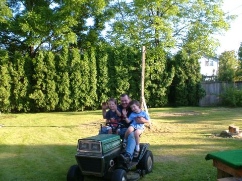 1989 Ford Bronco - A man and two children wave from a green lawn tractor in a backyard setting.