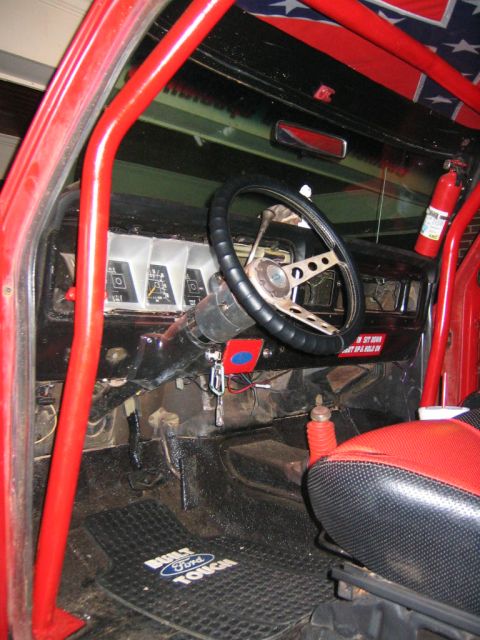 1979 Ford Bronco - Stripped interior of an older Ford 4x4 seen through the open driver's door, with a red-painted roll cage, black dash, three-spoke steering…