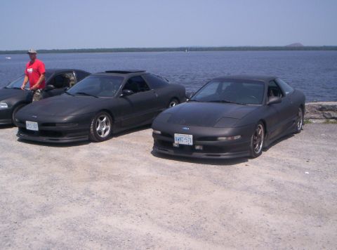 1993 Ford Probe - Two matte black 1993 Ford Probes are parked near the water, with a person standing by one of them.