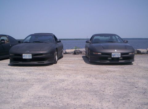 1993 Ford Probe - Two Ford Probes are parked side-by-side on a gravel lot near a body of water under a clear sky.