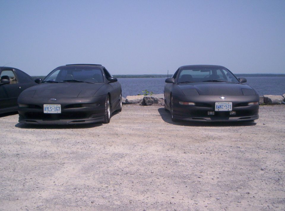 Two matte black Ford Probe coupes parked on gravel by water, front view