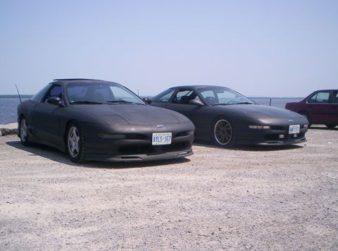 1993 Ford Probe - Two matte black Ford Probes are parked side-by-side on a gravel surface with a body of water in the background.