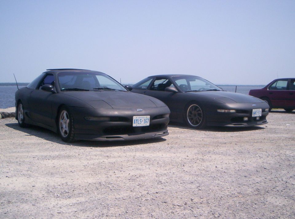 Two matte-black Ford Probe coupes parked on gravel by water